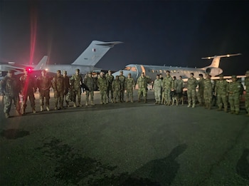 Grupo de soldados uniformados con un perro militar en una pista de aterrizaje de noche, con dos aviones al fondo