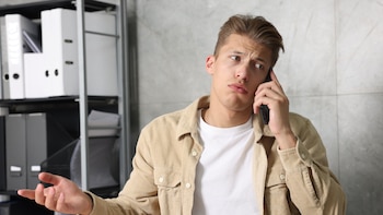Un hombre joven habla por teléfono mientras muestra una expresión de queja, en lo que parece ser una oficina.