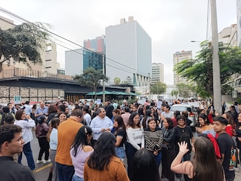 Vista general de una multitud de personas en una calle urbana con edificios altos y vehículos estacionados bajo un cielo nublado