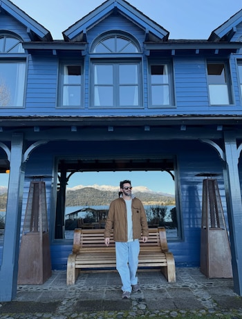 Hombre con campera marrón y pantalón claro. De pie frente a una casa azul. Los ventanales reflejan montañas y un lago. Un banco y dos calentadores