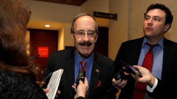 Un hombre con bigote y gafas habla mientras un periodista graba. A su derecha, otro hombre joven observa y sostiene un teléfono móvil