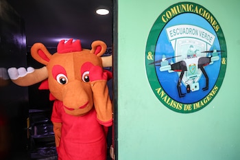 Una persona con un disfraz de mascota con cuernos marrones y traje rojo asoma por una puerta oscura, junto a un logo verde y azul del 'Escuadrón Verde'