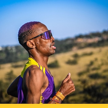 Hombre atleta con cabello morado, gafas de sol, camiseta deportiva púrpura y amarilla, pulsera y brazaletes de cuentas en entorno natural con cielo azul