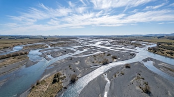Fotografía aérea gran angular de un río trenzado en Canterbury, mostrando múltiples canales de agua azul claro entre extensos bancos de grava gris y un cielo azul con nubes.