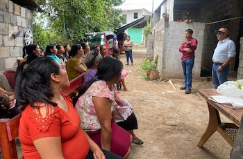 Grupo de personas, mayormente mujeres y niños, posando al aire libre con árboles al fondo, sosteniendo carteles blancos que mencionan programas agrícolas