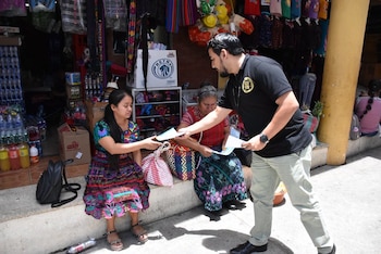 Un hombre de negro entrega papeles a dos mujeres con ropa tradicional sentadas en el escalón de una tienda con productos