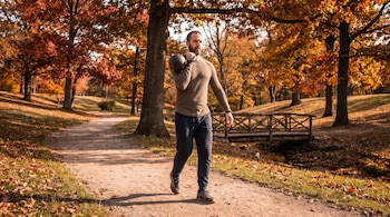 Hombre barbudo con camiseta térmica y pantalones deportivos lleva una pesa rusa negra en un parque con árboles de follaje otoñal, sendero de tierra y puente de madera sobre un arroyo.