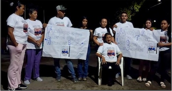 Un grupo de personas, incluyendo una mujer sentada, sostienen pancartas y usan camisetas blancas con la imagen de dos hombres, durante una protesta nocturna