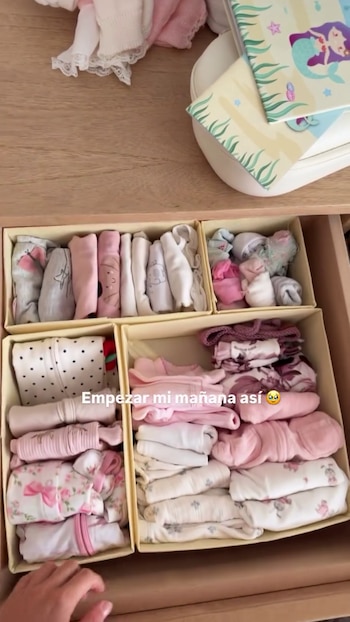 Un cajón abierto muestra ropa de bebé doblada y organizada en cuatro compartimentos. Las prendas son en tonos rosa, blanco y gris. Una mano toca el borde