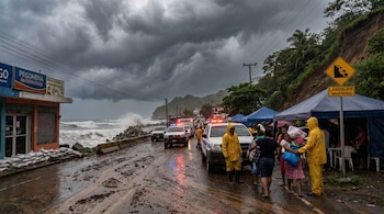 Carretera costera embarrada en El Salvador con cielo oscuro y olas grandes. Vehículos de emergencia, personas con impermeables y cartel de "Advertencia de Deslaves".