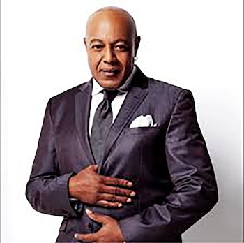 Peabo Bryson, un hombre calvo afroamericano, viste un traje oscuro con camisa blanca y corbata negra, posando con las manos cruzadas sobre el abdomen