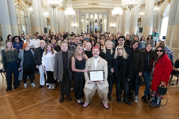 Un hombre con traje beige y gorro rojo sostiene un certificado en un gran salón con columnas y candelabros, rodeado por un grupo diverso de personas