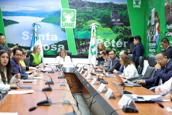 Personas sentadas en una mesa de conferencia con micrófonos, portátiles y banderas de Guatemala de fondo con logotipos del partido UNE