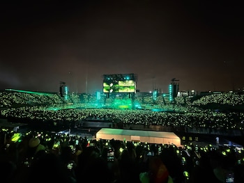 Multitud en un estadio nocturno iluminado por miles de luces verdes frente a un escenario central con pantallas