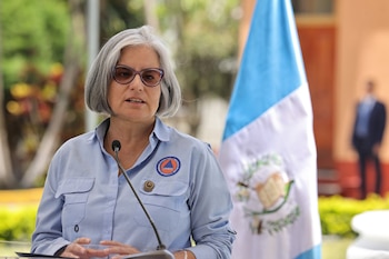 Mujer de cabello gris y gafas, con camisa azul y logo de CONRED, habla en un podio. Detrás, parcialmente visible, la bandera de Guatemala