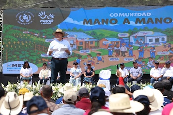 Un grupo de personas levanta la mano detrás de un cartel azul institucional en un terreno al aire libre con carpas de fondo