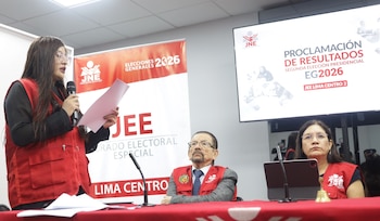 Mulher de óculos e vestido vermelho falando no microfone, segurando o papel. Duas pessoas com roupas vermelhas estão sentadas. Bandeira JNE. Tela com texto eleitoral
