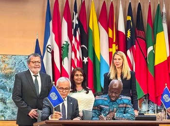 Gerardo Martínez habla desde un atril ante un fondo de banderas internacionales. Se ven arreglos de orquídeas blancas y el logotipo de la OIT en el escenario