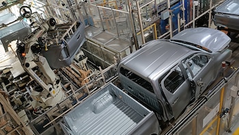 Vista aérea de una línea de ensamblaje de vehículos, mostrando robots industriales blancos y grises, y carrocerías de pickups plateadas sin terminar