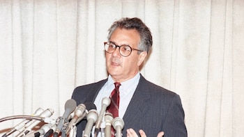 Retrato de Barney Frank, un hombre de mediana edad con gafas, traje a rayas oscuro y corbata burdeos, hablando detrás de varios micrófonos
