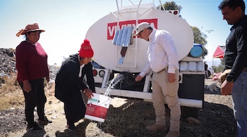 Un camión cisterna blanco vierte agua en un balde rojo y blanco. Una mujer sostiene el balde. Un hombre supervisa el flujo. Hay otras dos personas en el fondo