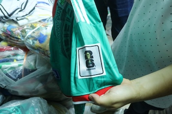 Primer plano de una persona sosteniendo una playera verde de fútbol con un parche que dice 'FIFA QUALIFIERS 2026'. Al fondo, pilas de mercancía en bolsas