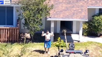Um homem sem toalha azul na cintura fica em frente a um prédio com as mãos para cima e um robô o observa.