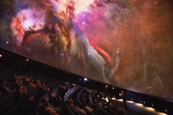 Una sala de planetario oscura con una gran proyección de una nebulosa de colores vibrantes en el techo abovedado y un grupo de personas sentadas