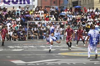 Jugador de fútbol en blanco y azul ejecuta una chilena con el balón en el aire, rodeado de otros jugadores y una gran multitud en un campo urbano