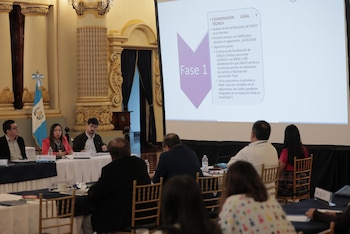 Un grupo de personas sentadas en mesas, observando una pantalla grande que muestra una presentación con texto y el título "Fase 1" en un salón con columnas
