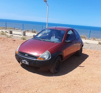Un coche subcompacto Ford Ka de color rojo y negro está estacionado sobre grava rojiza, con el océano azul y una valla azul detrás bajo un cielo despejado
