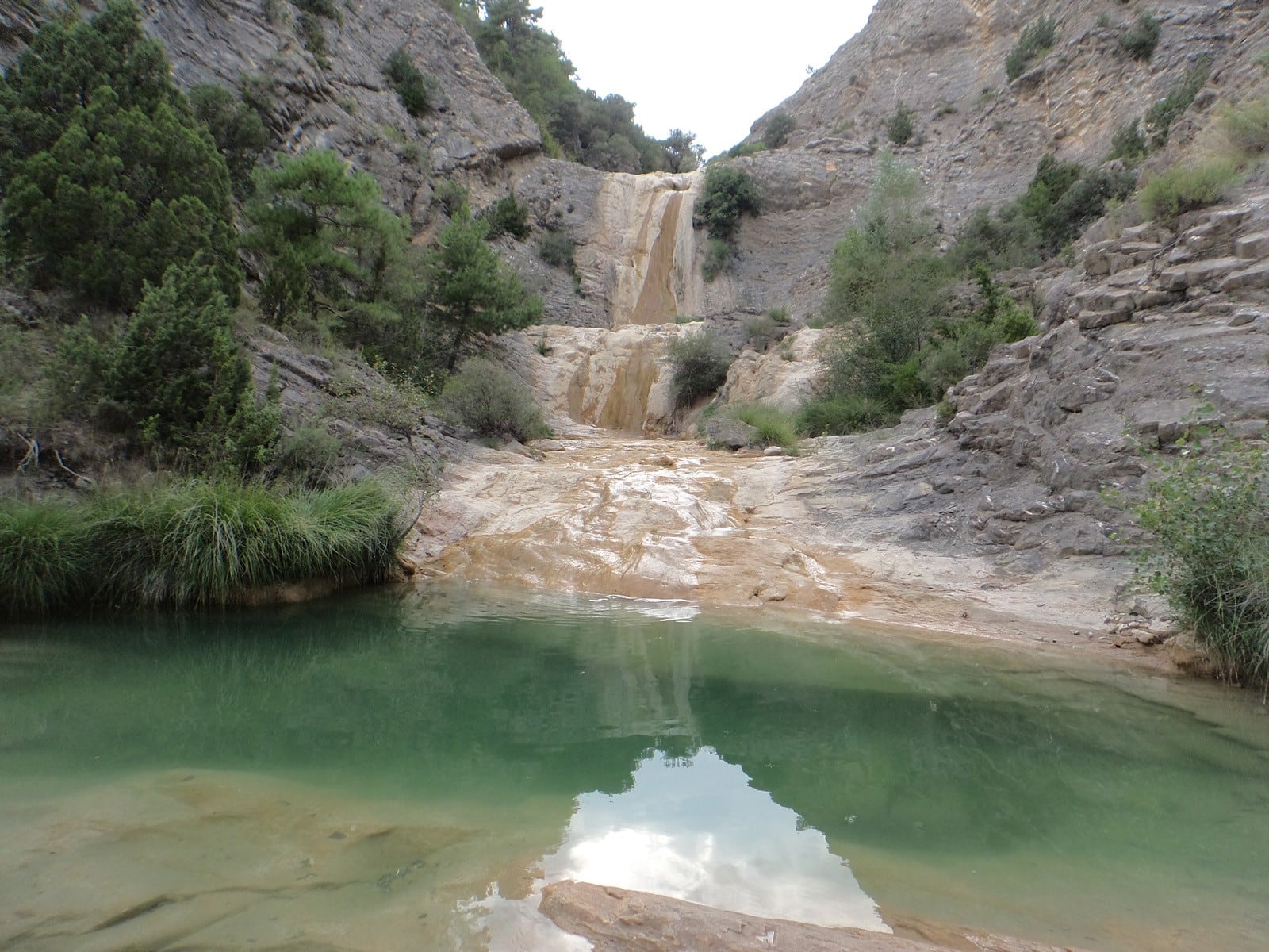 Una balsa de agua verdosa recibe la caída de un salto de agua entre paredes de roca y vegetación en las Balsas de Organyà