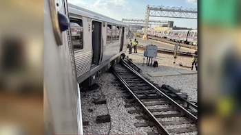 Un vagón de tren plateado aparece parcialmente descarrilado e inclinado sobre vías de grava. Varios trabajadores con chalecos reflectantes están cerca