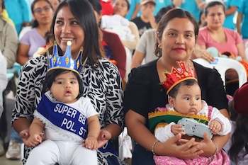 Dos mujeres sentadas sostienen en sus brazos a sendos bebés que llevan coronas decorativas y bandas distintivas