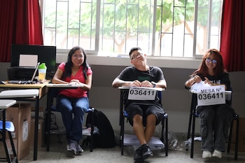 Tres personas sonrientes, dos mujeres y un hombre, sentadas en escritorios con letreros "MESA Nº 036411" en una sala luminosa