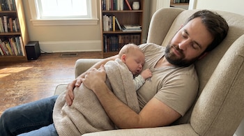 Un hombre con barba y un bebé duermen abrazados en un sillón beige, cubiertos con una manta clara, con estanterías y una ventana al fondo.