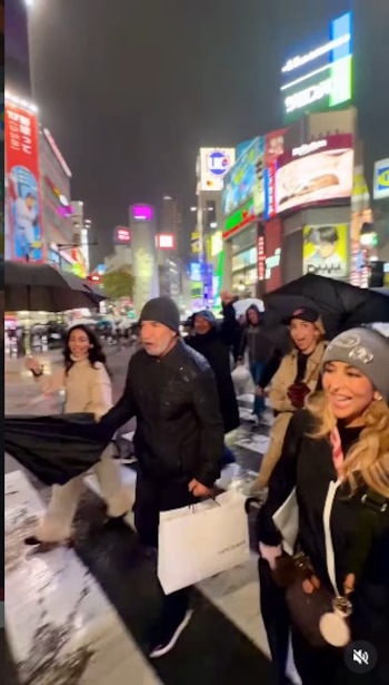 Un grupo de personas camina por un paso de cebra mojado en una ciudad iluminada de noche, con paraguas, edificios altos y carteles de neón visibles