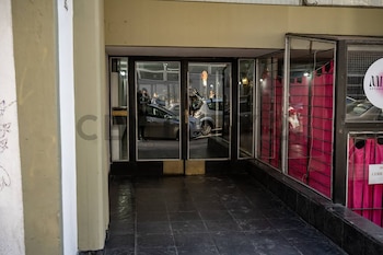 Entrada de un edificio con puertas de vidrio que reflejan coches y la calle, junto a una vitrina con cortinas rosas. El suelo es de baldosas oscuras