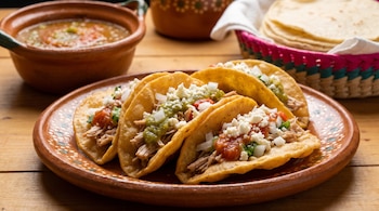 Cuatro chalupas servidas en un plato de barro con carne deshebrada, salsa, cebolla y queso, acompañadas de un recipiente con salsa y tortillas.