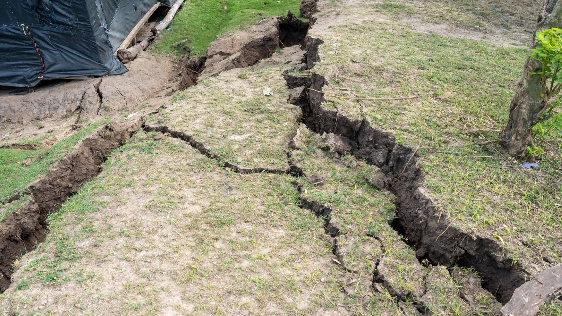 Extensas y profundas grietas en el suelo cubierto de pasto ralo son visibles tras un deslizamiento de tierra en la región San Martín, con una lona negra y troncos en el fondo. (Municipalidad El Dorado)