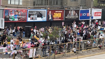 Multitud de personas en una calle junto a vallas metálicas, puestos de comercio ambulante y carteles publicitarios al fondo