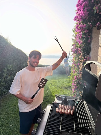 Hombre sonriente con espátula y tenedor de parrilla cocinando salchichas en una barbacoa. Detrás, una mujer de espaldas, césped, plantas con flores y un cuerpo de agua con botes
