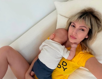 Primer plano de Úrsula Corberó acostada, con cabello rubio y camiseta amarilla, mirando a cámara mientras su bebé, de espaldas, descansa sobre su hombro