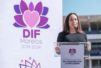 Una mujer habla frente a un micrófono sobre un atril transparente, al lado de un panel con el logotipo del DIF Morelos