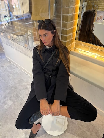 Lola Latorre sentada en un taburete blanco, vestida con gabardina y pantalones negros, gafas de sol en la cabeza, mirando a la izquierda en un local con paredes de ladrillo
