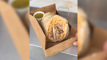 Un burrito grande y cortado por la mitad, mostrando arroz anaranjado, frijoles y carne, se encuentra en una bandeja de cartón junto a un pequeño recipiente con salsa verde; una mano sostiene la bandeja