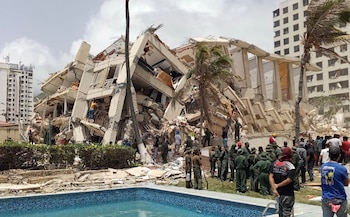 Edificio de hormigón colapsado con escombros y varillas expuestas, varias personas y militares alrededor, palmeras, piscina azul y otros edificios al fondo
