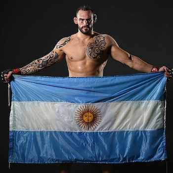 Hombre musculoso con tatuajes en brazos y pecho, barba, guantes de artes marciales mixtas, sosteniendo una bandera de Argentina