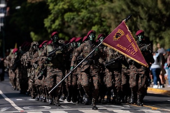 Grupo de soldados con uniformes camuflados, rostros pintados, boinas rojas y rifles, marchando. Un soldado lleva bandera burgundy con texto