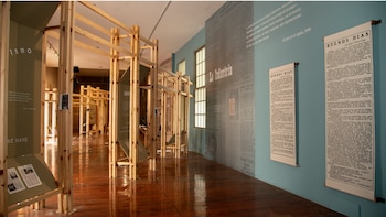 Sala de exposición con marcos de madera, paredes turquesas y verde oliva, y reproducciones de periódicos de gran tamaño. Dos pergaminos 'Buenos Días' cuelgan en la pared
